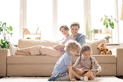 Family enjoying time together on a couch, with children using a tablet and a teddy bear nearby, illustrating indoor comfort and family bonding during changing seasons.