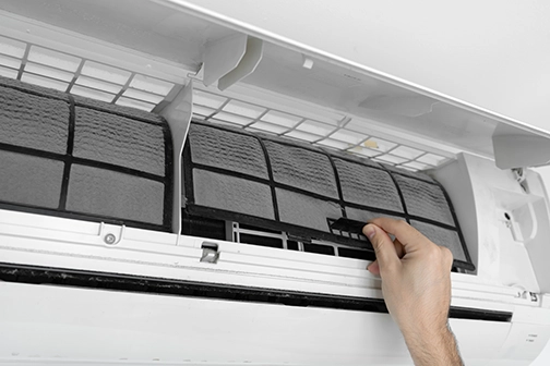 Hand removing a dirty HVAC filter from an air conditioning unit, emphasizing the importance of regular maintenance for energy efficiency and indoor air quality.